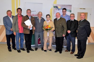 Eine Gruppe von Menschen, darunter eine ältere Frau mit Blumen, steht vor einem Banner. Sie alle lächeln und scheinen für ein Foto zu posieren.