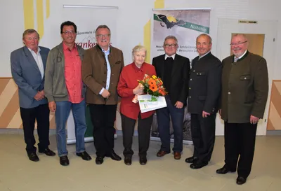 Eine Gruppe von Menschen steht zusammen, eine Frau in der Mitte hält einen Blumenstrauß und ein Zertifikat. Sie sind alle formell gekleidet. Hinter ihnen befindet sich ein Banner mit einer Fledermaus und Text.