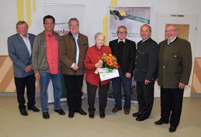 Eine Gruppe von Menschen steht zusammen, eine Frau in der Mitte hält einen Blumenstrauß und ein Zertifikat. Sie sind alle formell gekleidet. Hinter ihnen befindet sich ein Banner mit einer Fledermaus und Text.