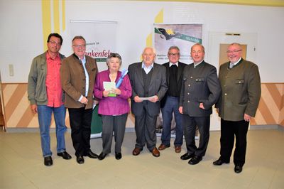 Eine Gruppe von Männern und einer Frau steht gemeinsam vor einem Banner und lächelt für ein Foto. Sie sind in formeller Kleidung gekleidet.