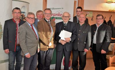 Eine Gruppe von Männern in formeller Kleidung, die vor einem Banner stehen, auf dem 'Willkommen in Ranke' steht. Der Mann in der Mitte hält ein Stück Papier.