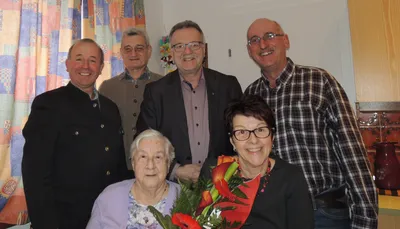 Fünf Menschen lächeln für ein Foto, wobei eine Frau einen Blumenstrauß hält. Hinter ihr sitzt eine ältere Frau mit einem Mann, der eine Brille trägt. Hinter ihnen stehen zwei Männer in Anzügen und ein Mann in einem karierten Hemd.