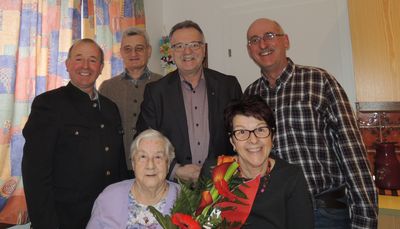 Fünf Menschen lächeln für ein Foto, wobei eine Frau einen Blumenstrauß hält. Hinter ihr sitzt eine ältere Frau mit einem Mann, der eine Brille trägt. Hinter ihnen stehen zwei Männer in Anzügen und ein Mann in einem karierten Hemd.