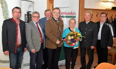 Fünf Männer und eine Frau stehen vor einem Banner mit dem Text 'Willkommen in Bankenfels'. Die Frau hält einen Blumenstrauß.