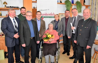 Eine Gruppe von Menschen steht um eine Frau im Rollstuhl, die Blumen hält, mit einem Schild im Hintergrund, das 'Willkommen in Frankenfels' steht.
