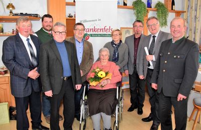 Eine Gruppe von Menschen steht um eine Frau im Rollstuhl, die Blumen hält, mit einem Schild im Hintergrund, das 'Willkommen in Frankenfels' steht.