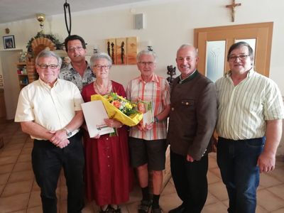 Eine Gruppe von sechs Erwachsenen, darunter eine Frau mit Blumen und einem Zertifikat, stehen in einem Raum. Sie alle lächeln.