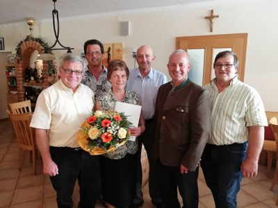Fünf Personen stehen lächelnd in einem Raum für ein Foto. Eine Frau hält einen Blumenstrauß und ein Stück Papier. Hinter ihnen sind ein Kreuz und eine Lampe an der Wand zu sehen.