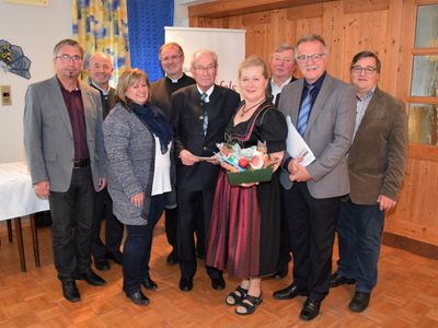 Eine Gruppe von Menschen, darunter Männer und Frauen, steht in einem Raum zusammen, möglicherweise für eine formelle Veranstaltung. Eine Frau in der Mitte hält einen Korb mit Früchten.