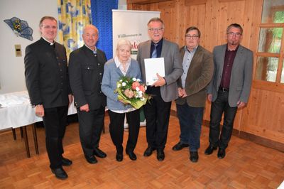Eine Gruppe von Männern und eine ältere Frau stehen lächelnd in einem Raum. Der Mann in der Mitte hält ein Zertifikat und einen Blumenstrauß. Hinter ihnen ist eine Holzwand mit einem Banner.