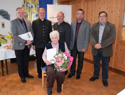 Eine ältere Frau mit einem Blumenstrauß wird von fünf Männern in formeller Kleidung umgeben, möglicherweise bei einer Zeremonie. Der Mann auf der linken Seite hält ein Stück Papier.