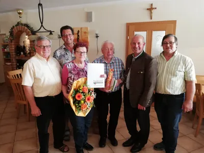 Eine Gruppe von Menschen steht lächelnd und posiert für ein Foto in einem Raum. Eine Frau hält einen Blumenstrauß und ein Zertifikat. Hinter ihnen hängen ein Kreuz und Bilderrahmen an der Wand.