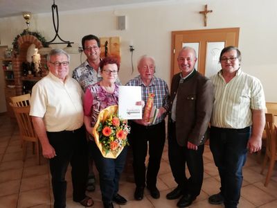 Eine Gruppe von Menschen steht lächelnd und posiert für ein Foto in einem Raum. Eine Frau hält einen Blumenstrauß und ein Zertifikat. Hinter ihnen hängen ein Kreuz und Bilderrahmen an der Wand.
