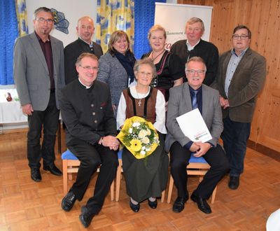 Eine Gruppe von Menschen, darunter eine Frau in traditioneller Kleidung, posiert für ein Foto in einem Raum mit Holzböden und einem Banner im Hintergrund.