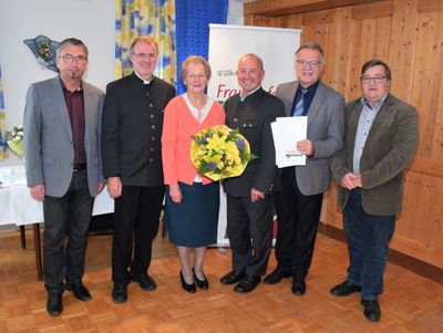 Eine Gruppe von sechs Personen, darunter eine Frau mit einem Blumenstrauß, steht für ein formelles Foto in einem Raum. Hinter ihnen befindet sich eine Holzwand mit einem Banner.