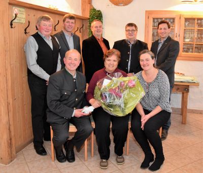 Eine Gruppe von Menschen in formeller Kleidung posiert für ein Foto mit einer Frau, die einen Blumenstrauß hält. Sie stehen in einem Raum mit Holzwänden und einem gefliesten Boden.
