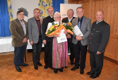 Eine Gruppe von Männern und einer Frau in traditioneller Kleidung halten Blumen und Urkunden in einem Raum.