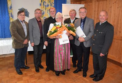Eine Gruppe von Männern und einer Frau in traditioneller Kleidung halten Blumen und Urkunden in einem Raum.