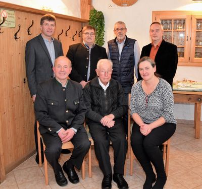 Eine Gruppe von sieben Personen, darunter Männer und Frauen, posiert für ein Foto in einem Raum mit Holzwänden und gefliestem Boden. Sie tragen alle formelle Kleidung.
