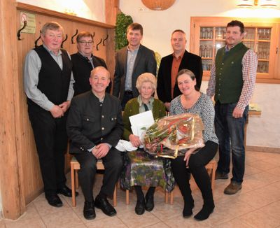 Eine Gruppe von Menschen steht und sitzt in einem Raum, eine Frau hält einen Blumenstrauß und ein Zertifikat.