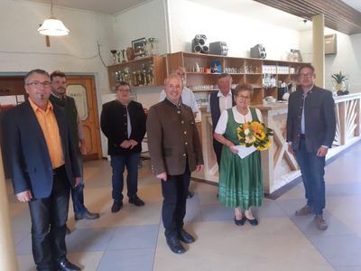 Eine Gruppe von Menschen steht in einem Raum, eine Frau hält einen Blumenstrauß. Hinter ihnen befindet sich ein Holzregal mit verschiedenen Gegenständen.