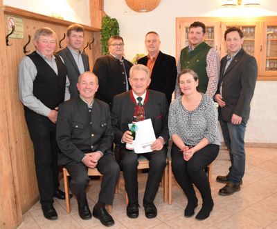 Eine Gruppe von Menschen, darunter Männer und Frauen, posiert für ein Foto in einem Raum. Ein Mann hält eine Flasche und ein Stück Papier. Sie alle lächeln und scheinen ein Ereignis zu feiern.