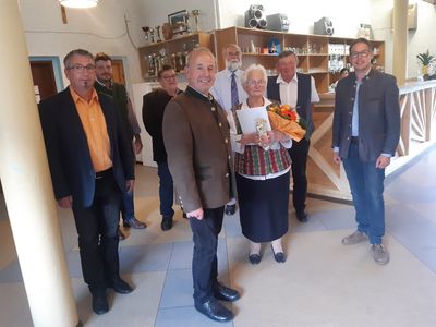 Eine Gruppe von Menschen steht in einem Raum, eine ältere Frau hält einen Blumenstrauß und eine Auszeichnung, ein Mann neben ihr lächelt und andere stehen hinter ihnen in der Nähe einer Theke.