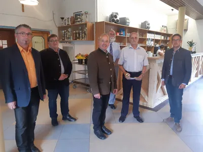 Eine Gruppe von Männern in formeller Kleidung steht in einem Raum mit einem Holztresen und Regalen, die mit verschiedenen Gegenständen gefüllt sind. Ein Mann hält einen Teller mit Essen.