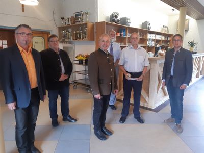 Eine Gruppe von Männern in formeller Kleidung steht in einem Raum mit einem Holztresen und Regalen, die mit verschiedenen Gegenständen gefüllt sind. Ein Mann hält einen Teller mit Essen.