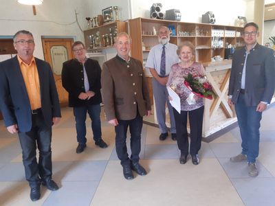 Eine Gruppe von Menschen steht in einem Raum mit einer Frau, die einen Blumenstrauß hält. Dahinter befindet sich eine Holztheke mit Regalen, die mit Gläsern und anderen Gegenständen gefüllt sind.