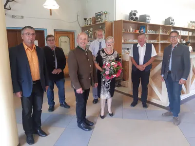 Eine Gruppe von Menschen steht in einem Raum, eine Frau in der Mitte hält einen Blumenstrauß. Hinter ihnen stehen Regale mit verschiedenen Gegenständen und eine Lampe hängt von der Decke.