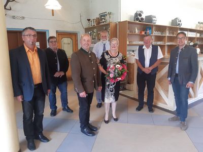 Eine Gruppe von Menschen steht in einem Raum, eine Frau in der Mitte hält einen Blumenstrauß. Hinter ihnen stehen Regale mit verschiedenen Gegenständen und eine Lampe hängt von der Decke.