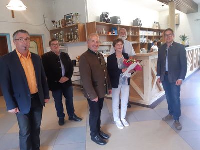 Eine Gruppe von Menschen steht in einem Raum mit einem Holztresen, Regalen und einer Person, die Blumen hält. Sie alle lächeln und scheinen für ein Foto zu posieren.