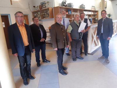 Eine Gruppe von Männern in formeller Kleidung steht in einem Raum und lächelt für ein Foto. Hinter ihnen sind Regale mit verschiedenen Gegenständen zu sehen, und eine Theke mit einem Blumenvase ist im Hintergrund.