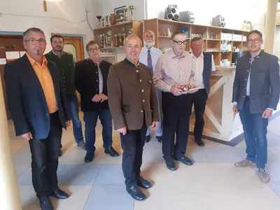 Eine Gruppe von Männern in formeller Kleidung steht in einem Raum mit Holzregalen und einer Theke im Hintergrund. Ein Mann hält eine Trophäe.