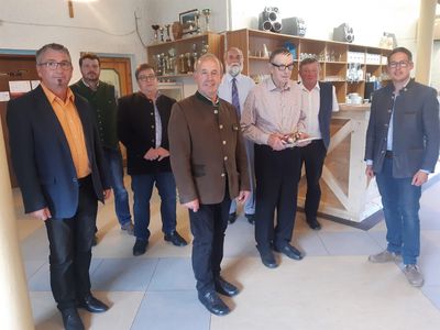 Eine Gruppe von Männern in formeller Kleidung steht in einem Raum mit Holzregalen und einer Theke im Hintergrund. Ein Mann hält eine Trophäe.