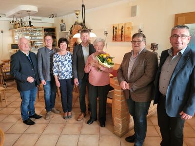 Eine Gruppe von Menschen steht lächelnd für ein Foto in einem Raum zusammen. Eine Frau hält einen Blumenstrauß.