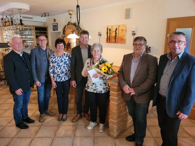 Eine Gruppe von Menschen steht in einem Raum zusammen, eine Frau hält einen Blumenstrauß und ein Zertifikat. Sie sind formell gekleidet und scheinen ein Ereignis zu feiern.