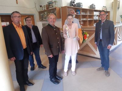 Eine Gruppe von Menschen steht in einem Raum, eine Frau hält eine Trophäe und lächelt. Hinter ihr steht ein Mann in einem braunen Jackett und auf der linken Seite ein Mann in einem orangefarbenen Hemd. Im Hintergrund befindet sich ein hölzerne Regal mit verschiedenen Gegenständen.
