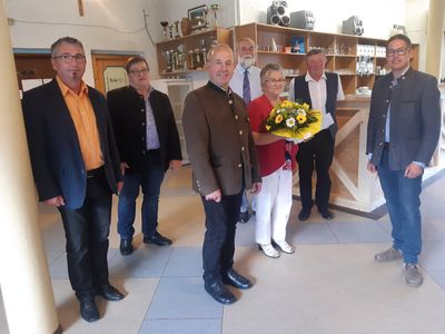 Eine Gruppe von Menschen steht in einem Raum, eine Person hält einen Blumenstrauß. Hinter ihnen befinden sich Regale mit verschiedenen Gegenständen.
