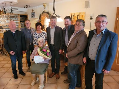 Eine Gruppe von Menschen steht und sitzt in einem Raum, eine ältere Frau hält Blumen und ein Papier, und ein Mann steht vor einer Ziegelwand.