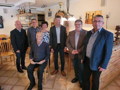 Eine Gruppe von sechs Personen, darunter ein älterer Mann im Stuhl, posiert für ein Foto in einem Raum mit gefliesten Böden und einem Holztisch.
