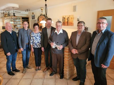 Eine Gruppe von Menschen, darunter Männer und Frauen, steht in einem Raum mit Ziegelwänden und einer Holztür. Ein Mann hält eine Flasche und Papier, während andere Brillen und Schuhe tragen.