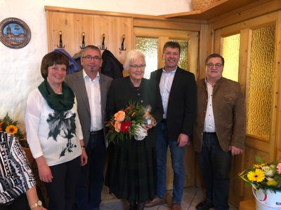 Fünf Menschen stehen lächelnd für ein Foto in einem Raum. Eine Frau in der Mitte hält einen Blumenstrauß. Hinter ihnen sind Holztüren und ein Kleiderständer mit Jacken zu sehen.