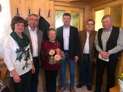 Fünf Menschen stehen in einem Raum und lächeln für ein Foto. Eine Frau hält einen Blumenstrauß. Hinter ihnen sind Holzwände mit Glasfenstern.