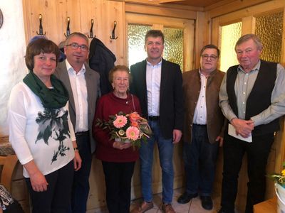 Fünf Menschen stehen in einem Raum und lächeln für ein Foto. Eine Frau hält einen Blumenstrauß. Hinter ihnen sind Holzwände mit Glasfenstern.