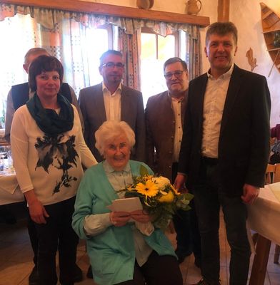 Eine ältere Frau mit einem Blumenstrauß wird von fünf anderen Personen umgeben, wahrscheinlich bei einer Feier.