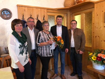 Fünf Menschen stehen lächelnd für ein Foto in einem Raum. Eine Frau hält einen Blumenstrauß. Hinter ihnen sind Holzwände und Glastüren zu sehen.