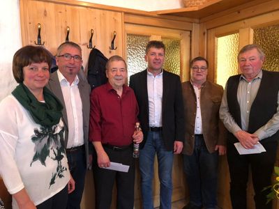 Fünf Menschen stehen lächelnd in einem Raum für ein Foto. Der Mann in der Mitte hält eine Flasche. Hinter ihnen ist eine Holzwand mit Haken.