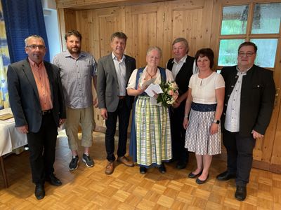 Eine Gruppe von Menschen steht in einem Raum mit Holzwänden und einem Holzboden und lächelt für ein Foto. Eine Frau in traditioneller Kleidung hält einen Blumenstrauß.